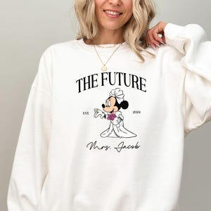 May include: White sweatshirt with a black graphic of a bride Mickey Mouse holding a ring. The text reads "The Future Est. 2024 Mrs. Jacob".
