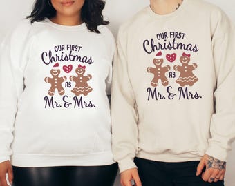 Op de afbeelding: Twee sweatshirts, één witte en één beige, met een kerstthema. Het ontwerp toont peperkoeken poppetjes en de tekst "Our First Christmas as Mr. & Mrs." in donkerpaars.