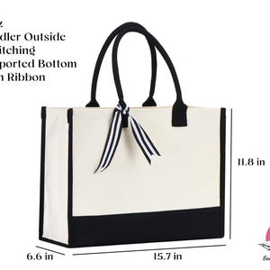 Op de afbeelding: Een witte canvas tote bag met zwarte bies en een zwarte bodem. De tas heeft een zwart lint dat om de handvatten is gebonden. De tas is 15,7 inch breed, 11,8 inch hoog en 6,6 inch diep.