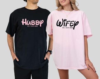 May include: A black t-shirt with the word "Hubby" in pink script and the date "Est. 14.07.2026". A light pink t-shirt with the word "Wifey" in black script and the date "Est. 14.07.2026".