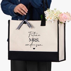 Op de afbeelding: Een witte canvas tote bag met zwarte afwerking en handvatten. De tas heeft een zwart lint om de handvatten gebonden en de tekst "Future Mrs. anderson" in zwart op de voorkant.