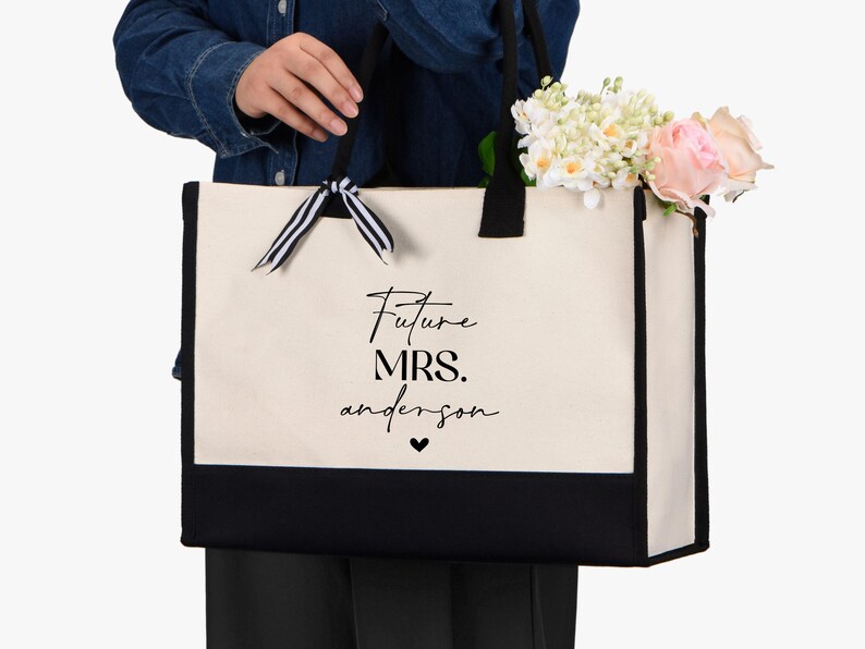 Op de afbeelding: Een witte canvas tote bag met zwarte afwerking en handvatten. De tas heeft een zwart lint om de handvatten gebonden en de tekst "Future Mrs. anderson" in zwart op de voorkant.