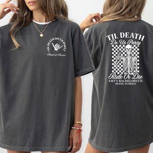 Może przedstawiać: Ciemnoszara koszulka z białym tekstem i grafiką. Prz&oacute;d koszulki m&oacute;wi "Til Death Do Us Party" z gestem dłoni i "Maid of Honor". Tył ma grafikę szkieletu oraz tekst "Ride or Die" i "Amy's Bachelorette Miami, Florida".