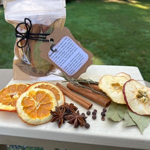 May include: A clear plastic pouch containing dried botanicals, tied with black string and a brown scalloped tag featuring text and a silver moon charm. Beside it are dried orange slices, cinnamon sticks, star anise, peppercorns, and apple slices.