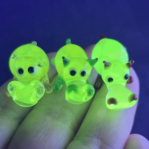 May include: Three small, neon green glass hippo figurines with black eyes and small ears. The hippos are in various poses, with some having small red accents. The figurines are held in a hand against a dark blue background.