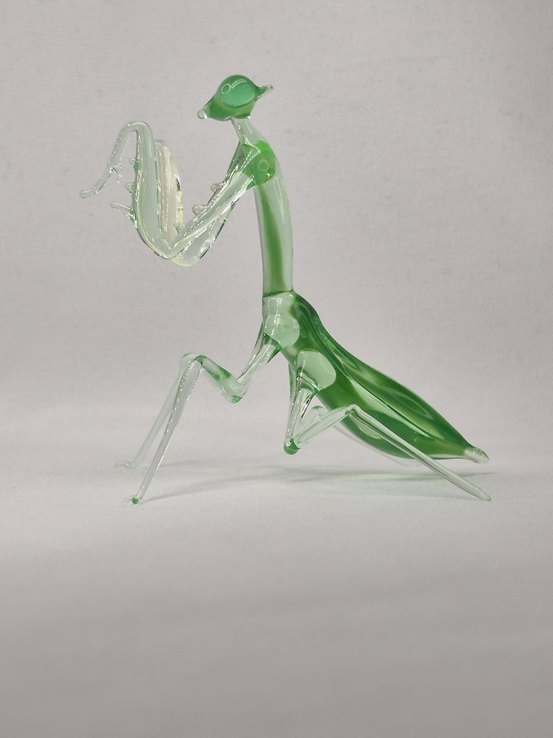 May include: A glass praying mantis sculpture. The insect is primarily clear with green accents on the head, back, and legs. The sculpture is detailed, with visible legs and antennae. The mantis is posed as if it is praying.