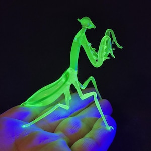 May include: A neon green glass praying mantis figurine. The insect is posed with its arms raised, and its body is elongated with detailed legs and antennae. The figurine glows brightly against a dark background.