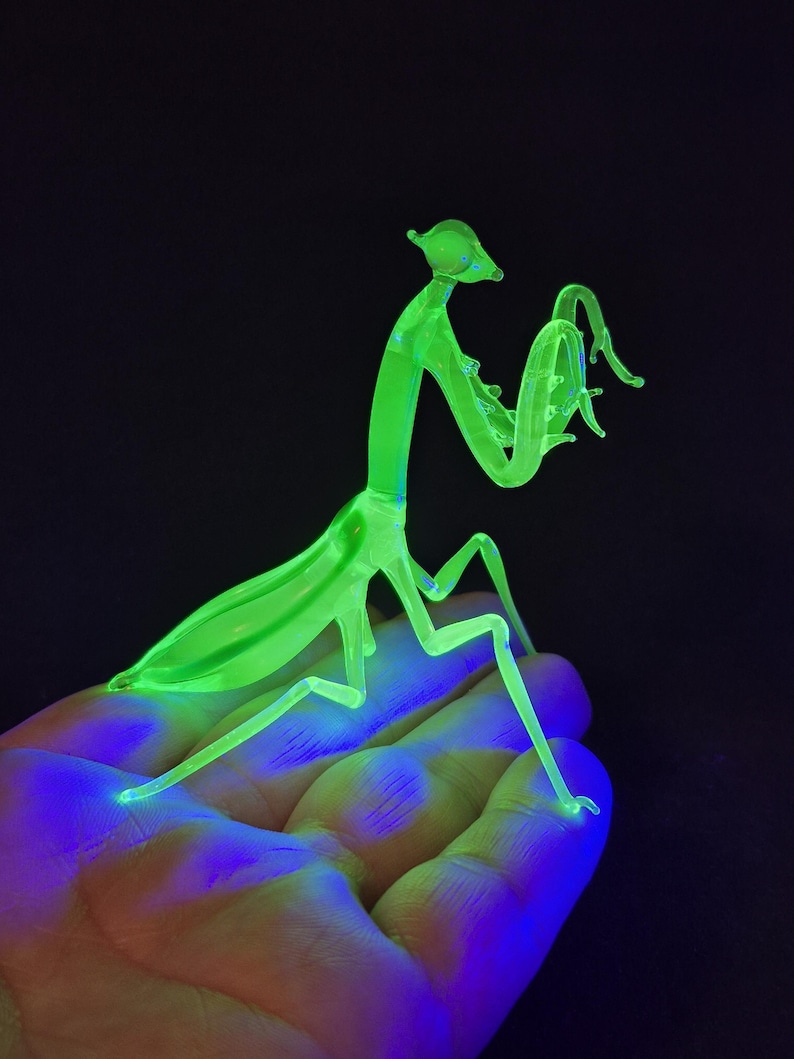 May include: A neon green glass praying mantis figurine. The insect is posed with its arms raised, and its body is elongated with detailed legs and antennae. The figurine glows brightly against a dark background.