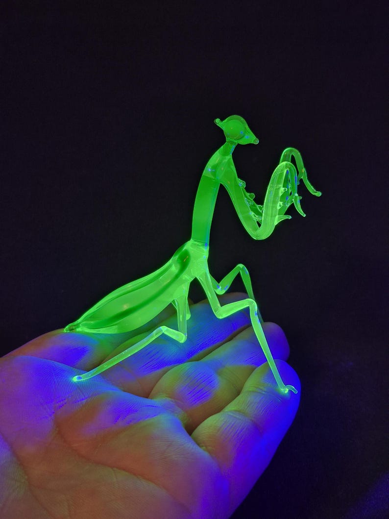 May include: A neon green glass praying mantis sculpture. The insect is posed with its front legs raised, and its body is illuminated against a dark background. The sculpture is a unique decorative item.