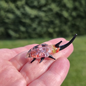 Puede incluir: Una pequeña y detallada figura de escarabajo de cristal. El escarabajo tiene un cuerpo translúcido con detalles rojos, rosas y negros. Presenta patas y antenas negras, y se sostiene en la mano de una persona sobre un fondo verde borroso.