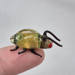 Op de afbeelding: Een kleine glazen insectenfiguur met een goudglitter lichaam en een heldere, groen getinte kop. Het insect heeft zwarte poten en antennes en rode accenten op de ogen. De figuur wordt door een vinger vastgehouden.