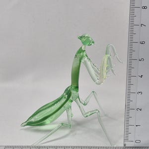 May include: A translucent green glass praying mantis figurine. The insect is depicted with detailed legs, antennae, and body segments. The figurine is approximately 6 inches long, with a ruler in the background for scale.