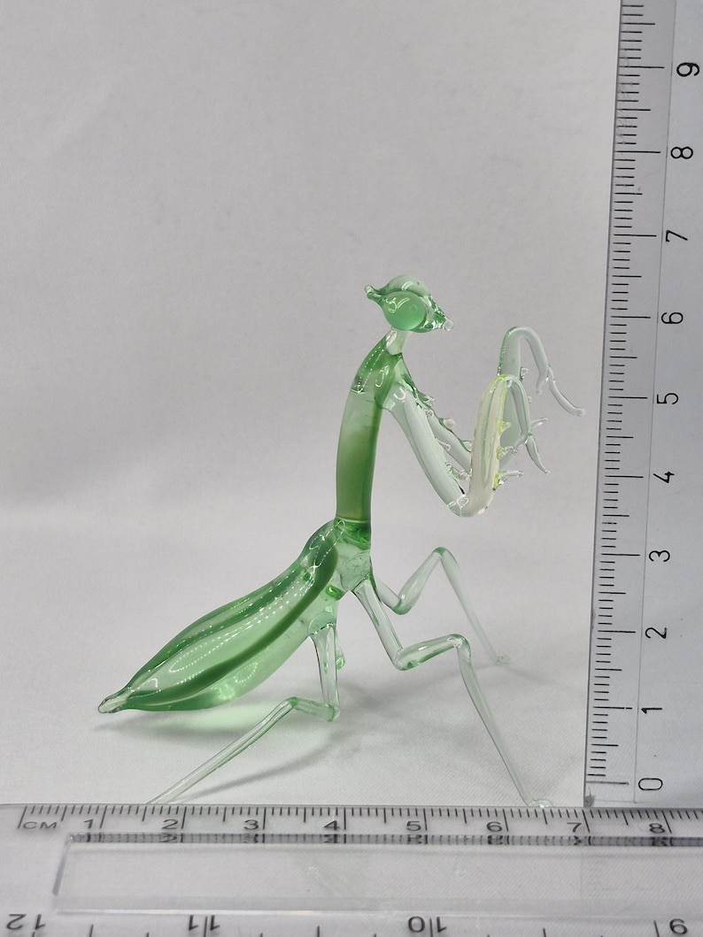 May include: A translucent green glass praying mantis figurine. The insect is depicted with detailed legs, antennae, and body segments. The figurine is approximately 6 inches long, with a ruler in the background for scale.