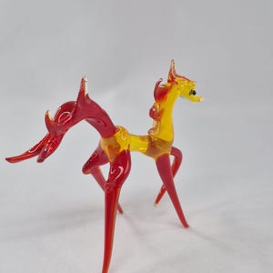 Może przedstawiać: Szklana figurka mitycznego stworzenia, w kolorach czerwonym i ż&oacute;łtym. Figurka ma smukłe ciało i długie, spiczaste nogi. Głowa i ogon zdobione są skomplikowanymi detalami.