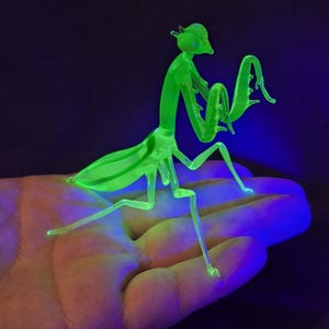 May include: A neon green praying mantis figurine with a detailed, insect-like form. The figurine is made of a translucent material, with visible legs, antennae, and wings. The mantis is posed on a surface, illuminated by a black light, creating a glowing effect.