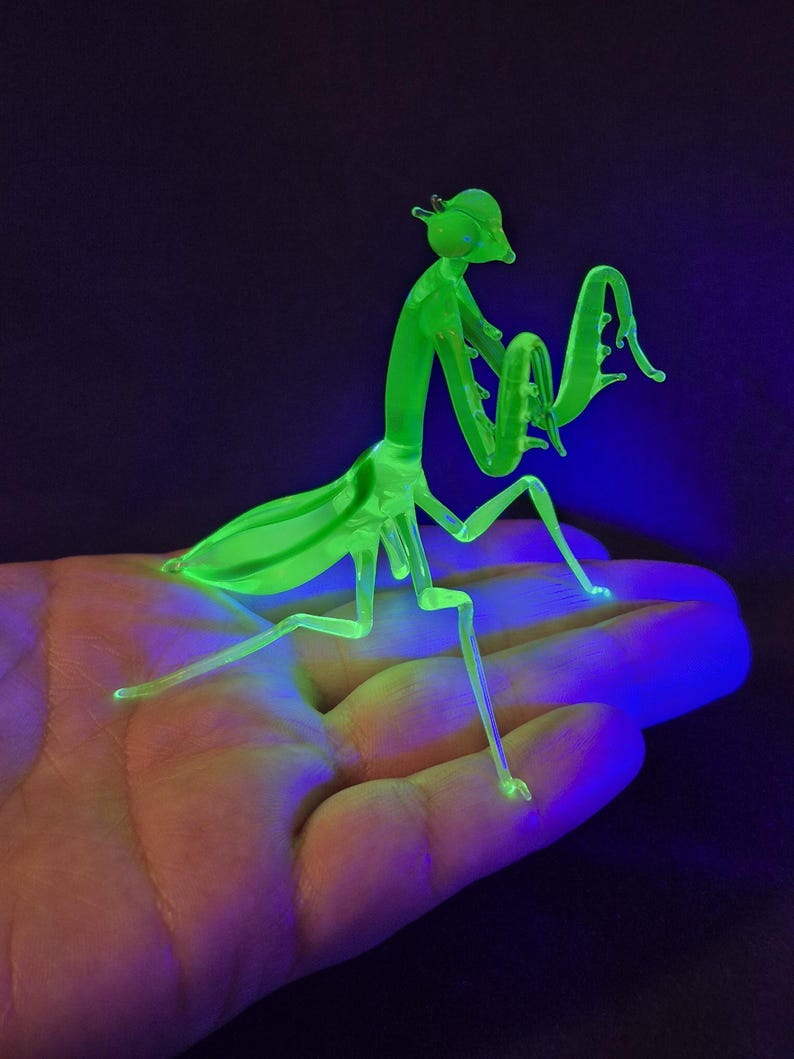 May include: A neon green praying mantis figurine with a detailed, insect-like form. The figurine is made of a translucent material, with visible legs, antennae, and wings. The mantis is posed on a surface, illuminated by a black light, creating a glowing effect.