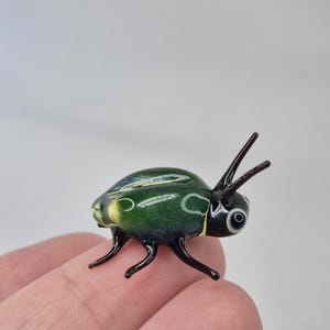 Puede incluir: Una figura de escarabajo de vidrio hecha a mano. El escarabajo es principalmente verde oscuro con patas y antenas negras. Tiene ojos blancos y negros y detalles amarillos. La figura se sostiene en la mano de una persona.