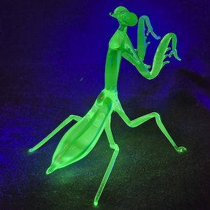 May include: A neon green glass praying mantis figurine. The insect sculpture has a slender body, long legs, and curved arms. The figurine glows brightly against a dark blue and black background.