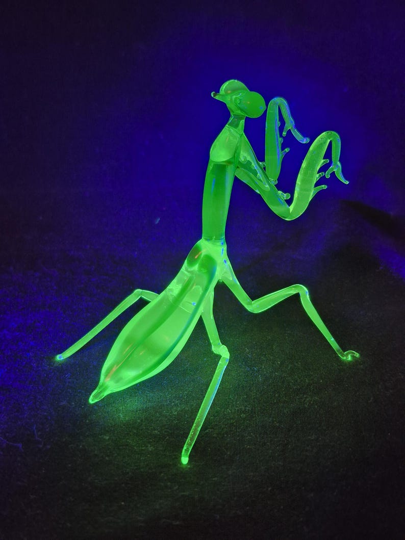May include: A neon green glass praying mantis figurine. The insect sculpture has a slender body, long legs, and curved arms. The figurine glows brightly against a dark blue and black background.