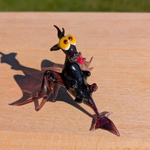 May include: A quirky glass ornament depicting a creature with large yellow eyes, a red heart-shaped detail, and purple legs and tail. The figurine is black and rests on a wooden surface.