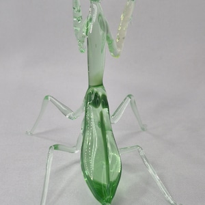 May include: A glass praying mantis figurine. The insect is primarily light green with clear legs and antennae. The figurine is posed with its arms raised, showcasing its delicate glass construction. The details include the insect's eyes and body segments.