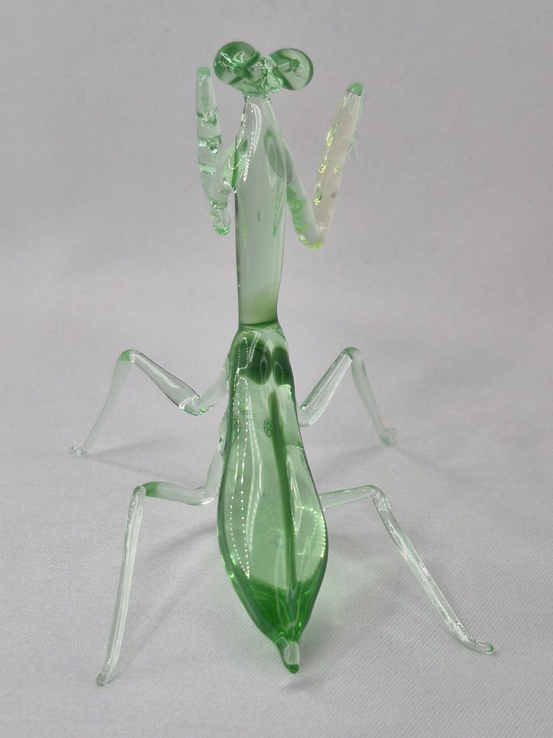 May include: A glass praying mantis figurine. The insect is primarily light green with clear legs and antennae. The figurine is posed with its arms raised, showcasing its delicate glass construction. The details include the insect's eyes and body segments.