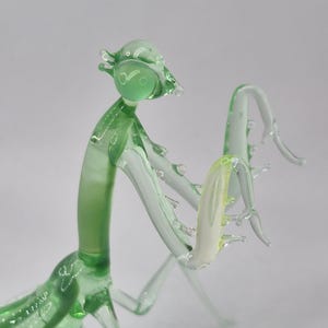 May include: A handcrafted glass praying mantis sculpture. The insect is primarily light green with clear accents, and a pale yellow section. The sculpture has a detailed, artistic design, with delicate legs and antennae. The glass is transparent, allowing light to pass through.