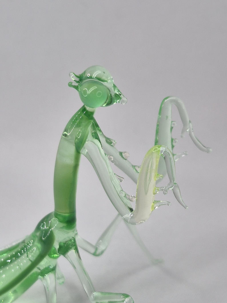 May include: A handcrafted glass praying mantis sculpture. The insect is primarily light green with clear accents, and a pale yellow section. The sculpture has a detailed, artistic design, with delicate legs and antennae. The glass is transparent, allowing light to pass through.