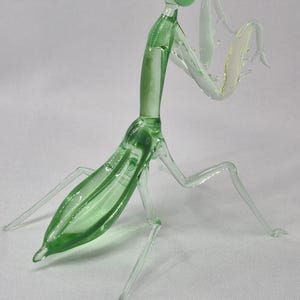 May include: A detailed glass sculpture of a praying mantis. The insect is primarily green with clear, delicate legs and arms. The sculpture has a smooth, glossy finish and is posed in a dynamic stance, showcasing its intricate details.