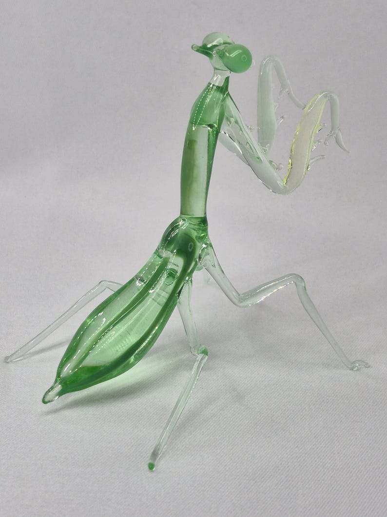May include: A detailed glass sculpture of a praying mantis. The insect is primarily green with clear, delicate legs and arms. The sculpture has a smooth, glossy finish and is posed in a dynamic stance, showcasing its intricate details.