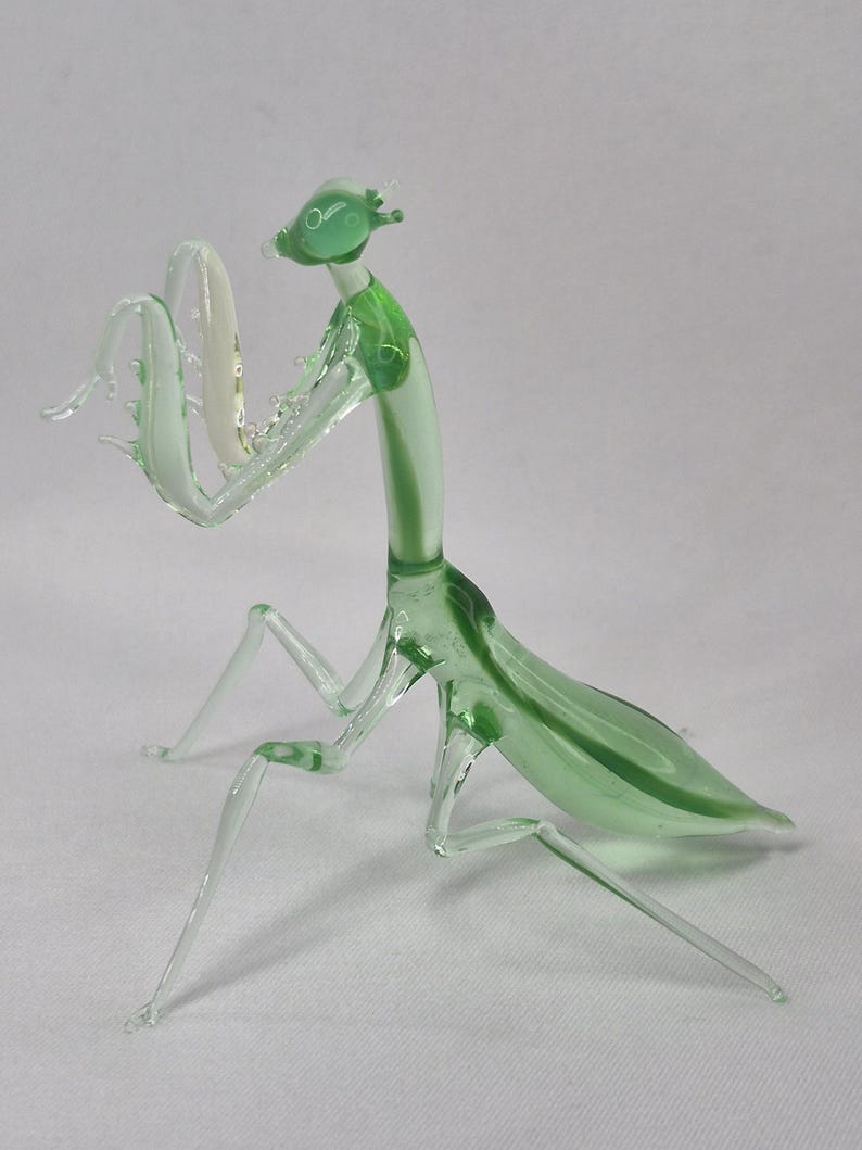 May include: A glass praying mantis figurine in shades of green and clear. The insect has a detailed head, long legs, and a curved body. The figurine is made of transparent glass, allowing light to pass through.