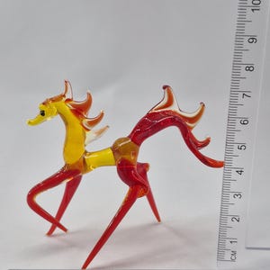 Może przedstawiać: Ręcznie wykonana szklana figurka konia w odcieniach ż&oacute;łci i czerwieni. Ozdobny koń ma około 10 cm wysokości, ze smukłym korpusem i stylizowaną grzywą i ogonem. Figurka jest umieszczona na białym tle.
