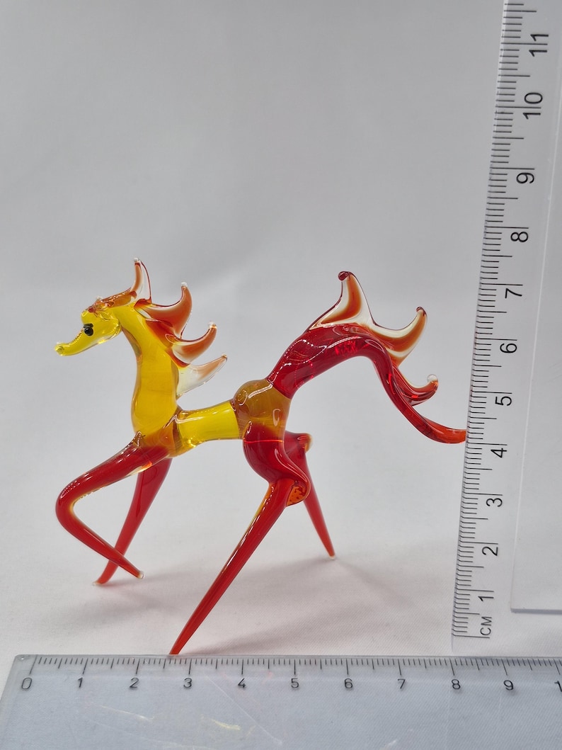 Może przedstawiać: Ręcznie wykonana szklana figurka konia w odcieniach ż&oacute;łci i czerwieni. Ozdobny koń ma około 10 cm wysokości, ze smukłym korpusem i stylizowaną grzywą i ogonem. Figurka jest umieszczona na białym tle.