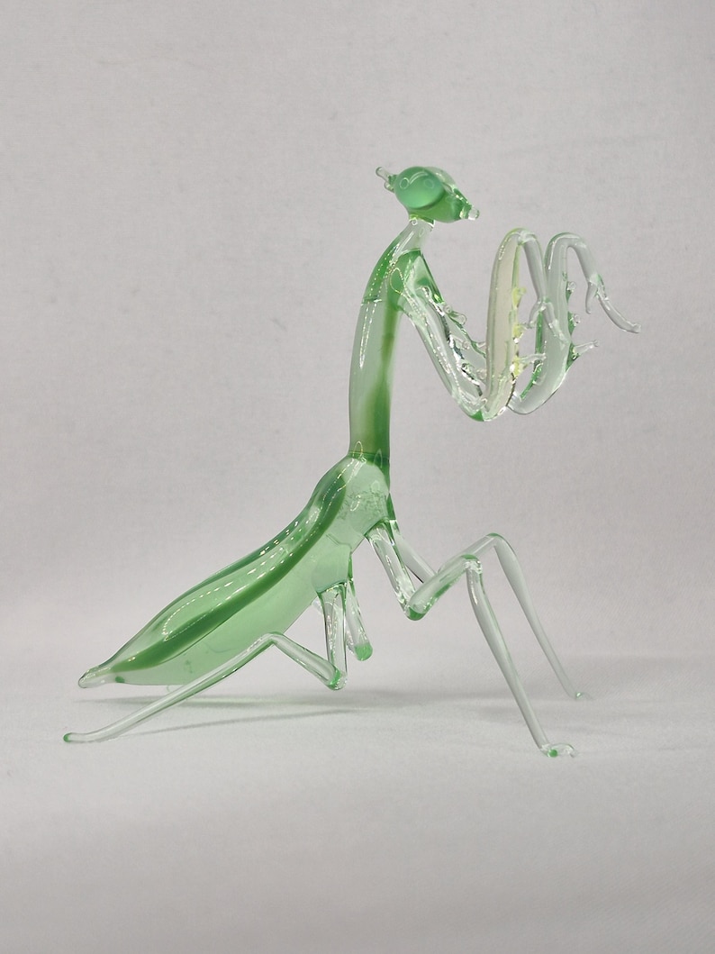 May include: A translucent green glass praying mantis figurine. The insect has a detailed body with outstretched arms and delicate legs. The figurine is a decorative item, showcasing the form of the insect.