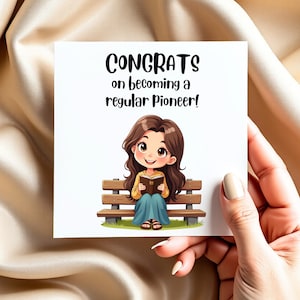 Puede incluir: Una tarjeta de felicitación blanca con el texto "CONGRATS on becoming a regular Pioneer!" presenta una ilustración de dibujos animados de una persona leyendo un libro mientras está sentada en un banco del parque. La persona tiene cabello castaño y usa una camisa amarilla y una falda azul.