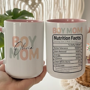 Könnte beinhalten: Zwei weiße Keramiktassen mit rosa Henkeln und Innenseiten. Eine Tasse zeigt den Text "BOY MOM" in gedeckten Farben mit dem Namen "Olivia" in schwarzer Schreibschrift. Die andere Tasse zeigt ein "Nutrition Facts"-Etikett mit humorvollem Text.