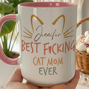 May include: White ceramic mug with a pink interior and handle. The mug features a cat silhouette and the text "Jennifer BEST FUCKING CAT MOM EVER" in various colors. A perfect gift for cat lovers.