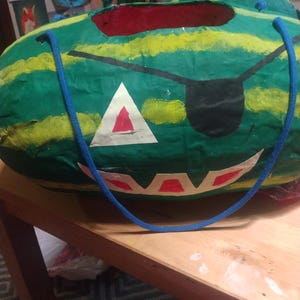 May include: A green and yellow watermelon-shaped papier-mâché pinata with a blue rope handle. The pinata has a red triangle, a black eye patch, and a white and red mouth.