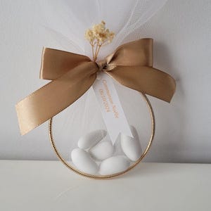 Puede incluir: Un recuerdo en forma de anillo dorado con tul blanco y un lazo de cinta dorada. Dentro del anillo hay caramelos blancos. Una pequeña etiqueta dice "Communion Noélie 06/10/2024". Flores secas están unidas al lazo.