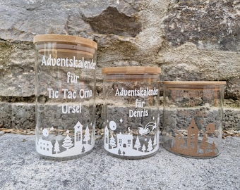 May include: Three glass jars with bamboo lids, each with white text and a winter village design. The text on the jars reads "Adventskalender für Tic Tac Oma Ursel", "Adventskalender für Dennis", and "Adventskalender".