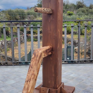 May include: A wooden martial arts training dummy with a brown stain finish. The dummy has three arms, one of which is angled downwards. The dummy is standing on a tiled patio.