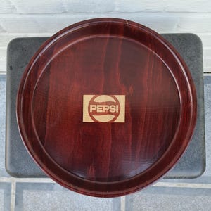 May include: A round wooden tray with a brown stain. The tray has a Pepsi logo in the centre.