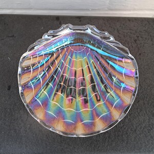 Op de afbeelding: Iriserende glazen schelpvormige schaal. De schaal heeft een geschulpte rand en een waaiervormig patroon met regenboogtinten van blauw, paars, geel en groen. De schaal is helder en reflecteert licht.