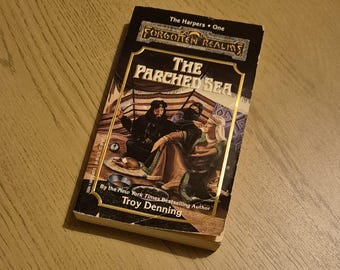 May include: A paperback book cover with a black background and gold accents. The title is "The Parched Sea" and the author is Troy Denning. The book is part of the Forgotten Realms series.