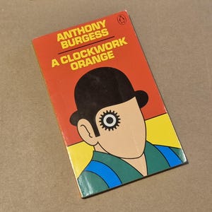 May include: A copy of Anthony Burgess's "A Clockwork Orange" paperback book. The cover is orange with the title in yellow. It features a graphic illustration of a man with a bowler hat and a gear for an eye.