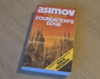Może przedstawiać: Książka w miękkiej okładce zatytułowana "Foundation's Edge" autorstwa Asimova. Okładka przedstawia ilustrację futurystycznego miasta na pomarańczowo-żółtym tle. Widoczny jest również tekst "The Fourth Book in the Epic Foundation Saga".