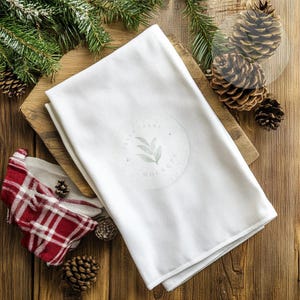 May include: A folded white kitchen towel with a circular logo featuring a leaf design is displayed on a wooden cutting board. A red and white plaid dish towel and pine cones are also visible, suggesting a holiday or rustic theme.