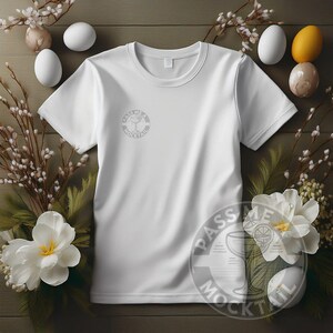 May include: White t-shirt with short sleeves and a round logo on the front that says "Pass Me A Mocktail". The shirt is lying on a wooden surface with white flowers and eggs.
