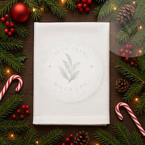 May include: A folded white cloth, possibly a towel or fabric, is the central focus, surrounded by Christmas decorations. Green pine branches, red berries, candy canes, a red ornament, and pine cones create a festive border. The cloth has a watermark that says "FRESH FRAME MOCKUPS".