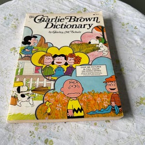 May include: The Charlie Brown Dictionary book cover features colorful illustrations of Charlie Brown, Snoopy, and other Peanuts characters. The book title is in bold, black letters with a red background. The text "Over 580 pictures in full color 2400 words defined" is printed in black on a white background.
